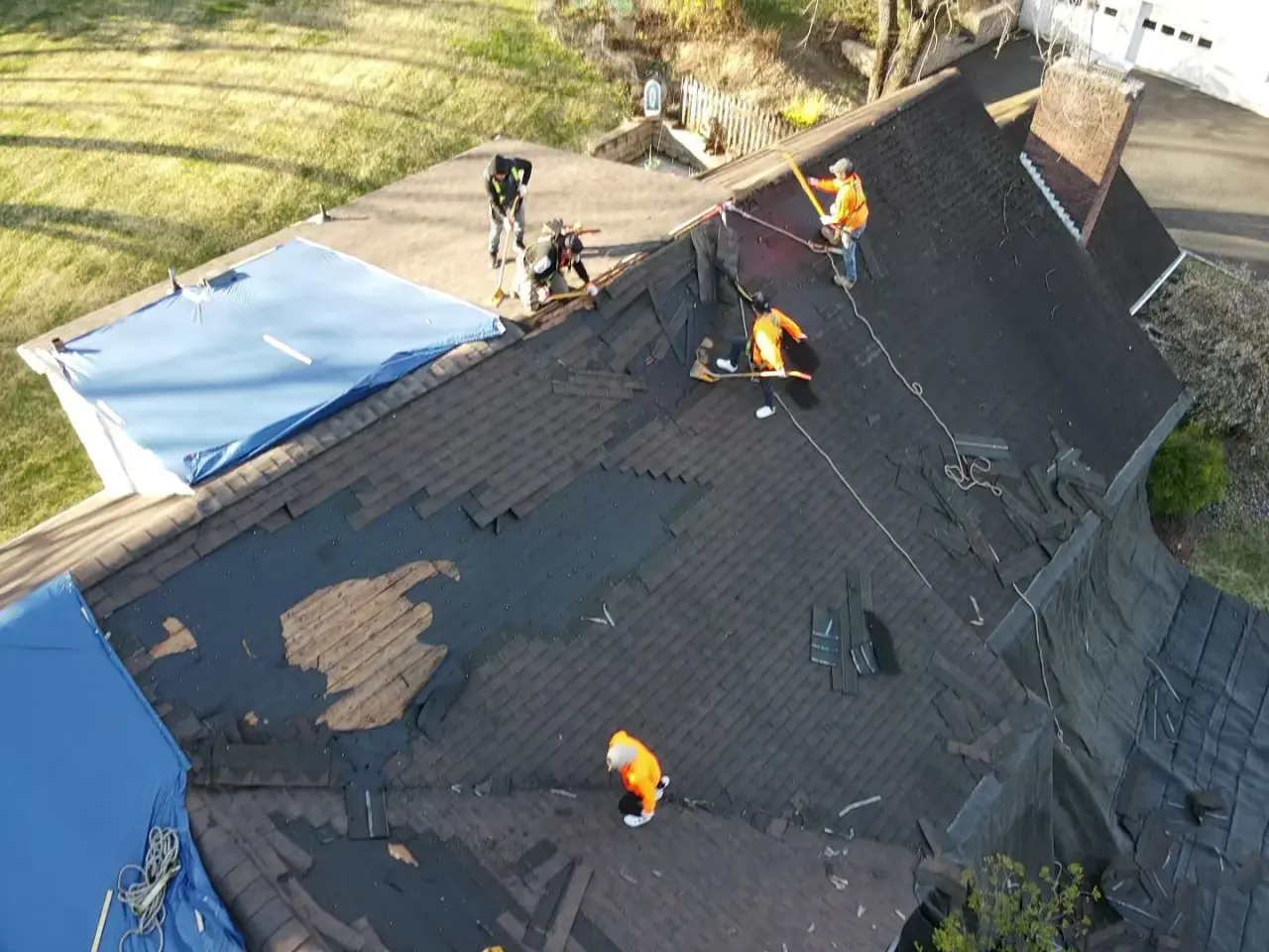 Roofers on a dark shingle roof with a damaged section opened up and tarps staged beside it