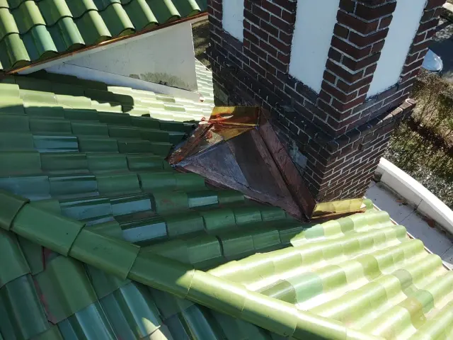 Copper cricket and flashing behind a brick chimney on a green clay tile roof