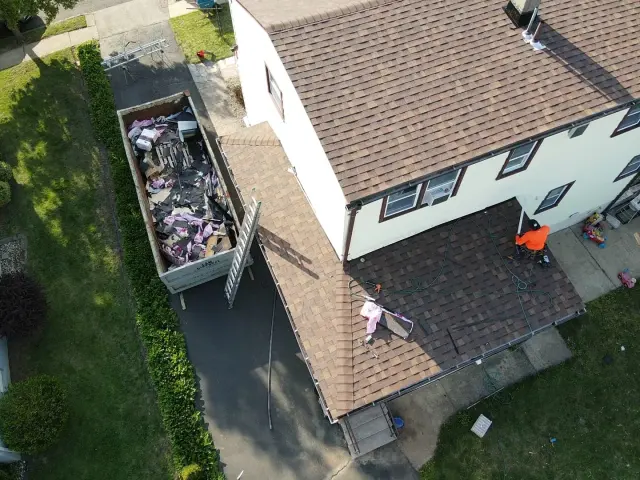 Roofer finishing the porch roof of a two-story home in new shingles, with the old roof in a dumpster below