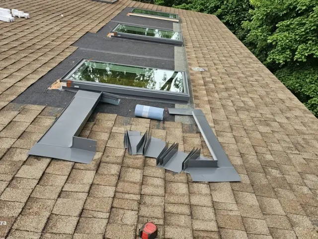 Skylight flashing kit pieces laid out on tan shingles beside the skylight opening