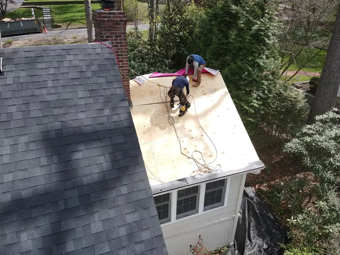 Two roofers laying underlayment over new plywood decking, with tarps protecting the garden below