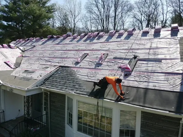 Roofer fastening shingles at the eave of a house surrounded by trees