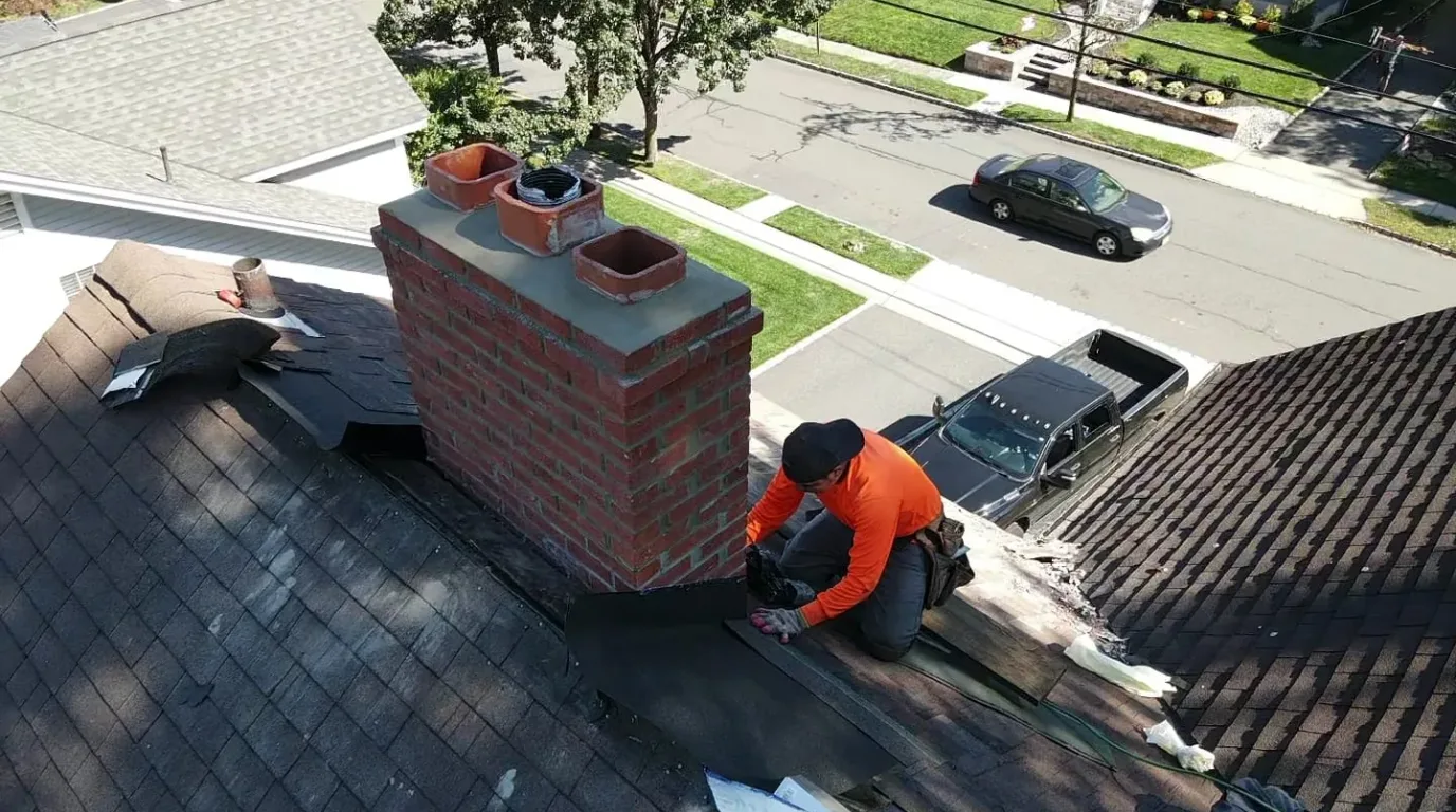 Roof leak repair in New Jersey: roofer setting new flashing membrane along the side of a brick chimney on a dark shingle roof